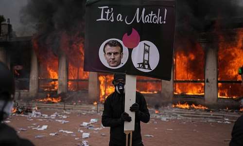A protester holds a placard with a photo of French president Emmanuel Macron and a guillotine in front of a burning tourism office during a demonstration against rising living costs and in solidarity with students grievances in Lyon on 17 October. - AP Photo/Laurent Cipriani - Laurent Cipriani