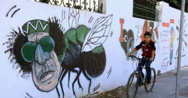 A Libyan boy cycles past graffiti depicting former strongman Moamer Kadhafi painted on a wall in Tripoli,. By Mahmud Turkia (AFP/File)