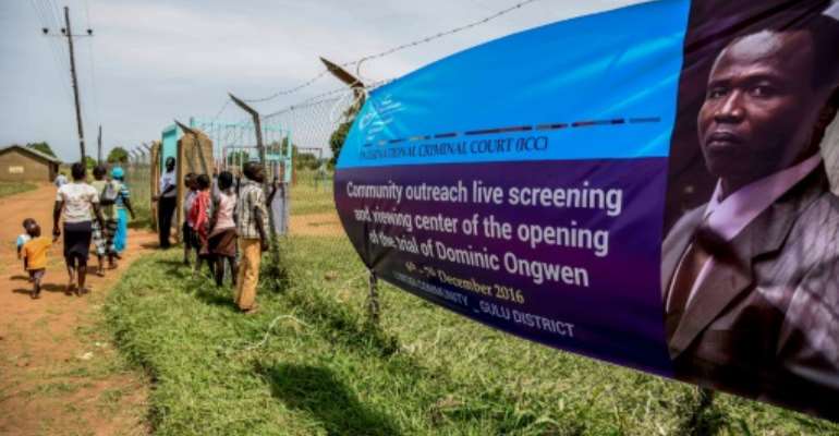 People walk near a banner of former child soldier-turned-warlord Dominic Ongwen before the screening of the start of his ICC (International Criminal Court) trial in Lukodi, Uganda on December 6, 2016. By ISAAC KASAMANI (AFP/File)