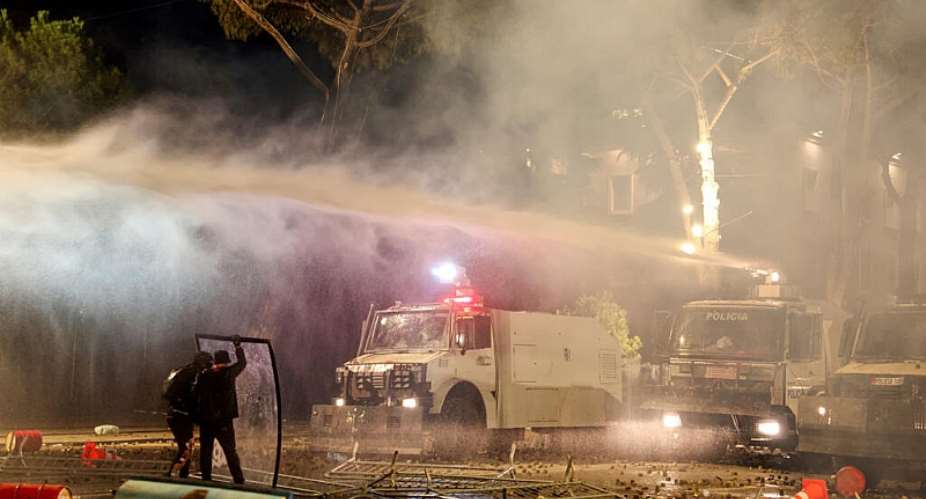 Protesters stand behind a makeshift shield as they face off with police water canon during an anti-government demonstration in central Tirana on October 16, 2026. -  Adnan Beci, AFP