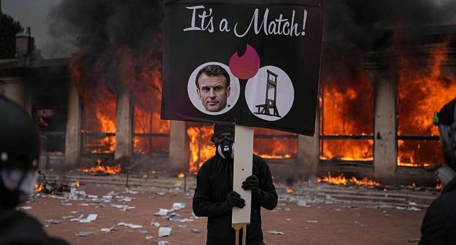 A protester holds a placard with a photo of French president Emmanuel Macron and a guillotine in front of a burning tourism office during a demonstration against rising living costs and in solidarity with students grievances in Lyon on 17 October. - AP Photo/Laurent Cipriani - Laurent Cipriani