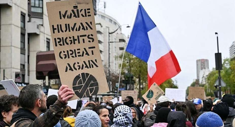 A protester holds a placard depicting a symbol in reference to Crass, an English art collective and anarcho-punk band, as another holds a French flag during a demonstration in favor of purchasing power, the protection of public services, and fiscal and social justice, in Paris on October 24, 2026 as part of a nationwide call for mobilisation bringing together former members of the Yellow Vest movement, labor unions, high school and university students. - AFP - NICOLAS TUCAT