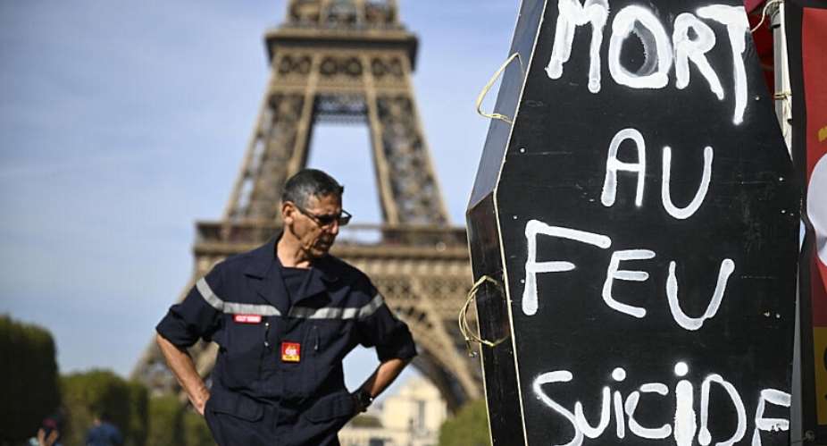 A firefighter stands next to a coffin-shaped sign reading Death by fire, suicide in front of the Eiffel Tower during a demonstration entitled the Reinforcement Column of Anger at the Champs de Mars in Paris, on 27 September, 2026. -  AFP - Julien de Rosa