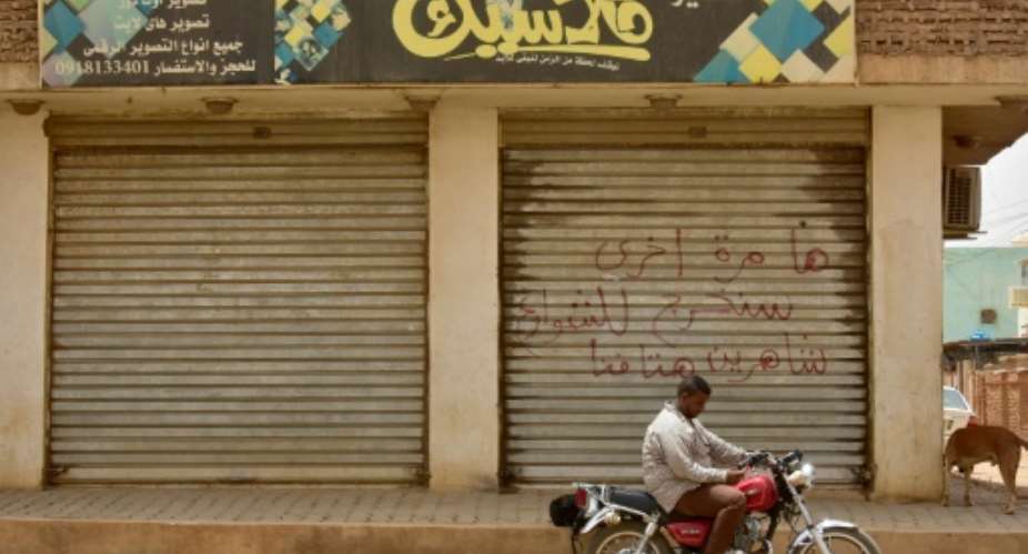 A Sudanese man sits on a motorbike in front of a closed shop in Khartoum with writing on its shutters that reads: Another time, we shall take to the streets, crying out our demands loudly.  By - AFP