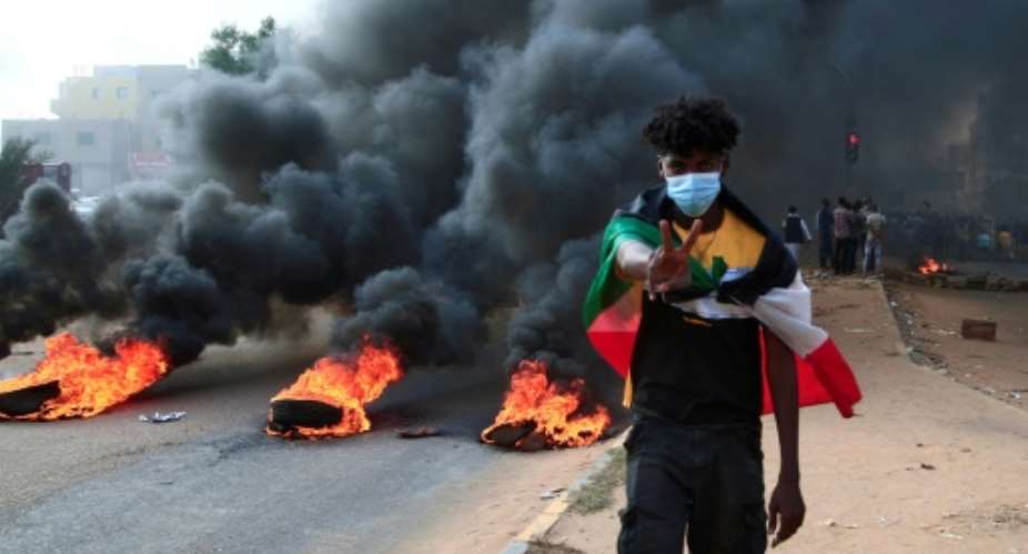 A Sudanese protester draped in the national flag flashes a V-for-victory sign next to burning tyres at a demonstration in Khartoum.  By - AFP