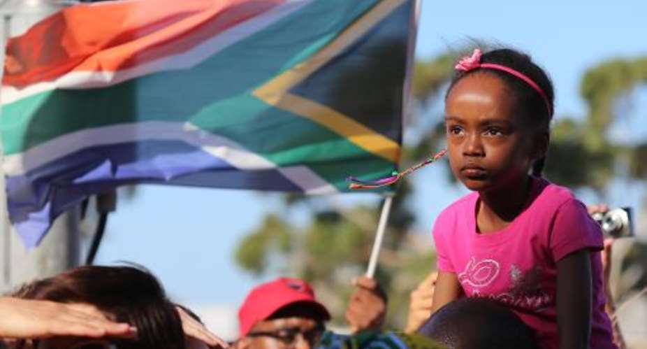 A girl looks on next to a man flying a South African flag as mourners gather to honour late South African former president Nelson Mandela at an inter-faith service on December 6, 2013, held at the Grand Parade in Cape Town.  By Jennifer Bruce AFP
