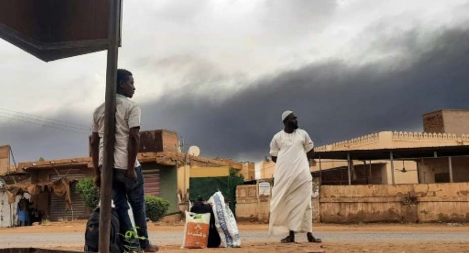 Smoke rises above buildings as people wait on the side of a road in Khartoum after a 24-hour ceasefire took effect.  By - (AFP)